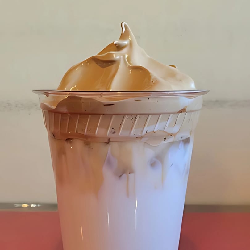 A close-up of a Dalgona coffee in a clear plastic cup, with whipped coffee foam on top.