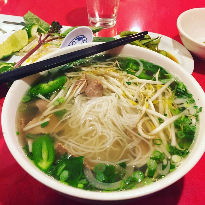 A bowl of Vietnamese pho with noodles, beef, bean sprouts, and green onions on a red table.