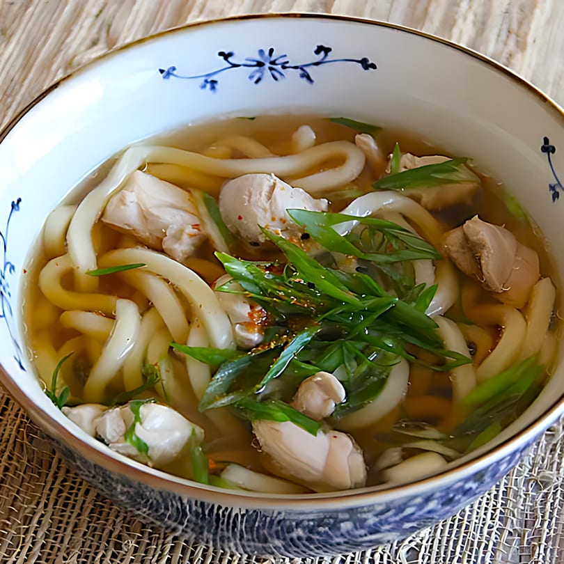 A bowl of Vietnamese chicken udon noodle soup, garnished with green onions and spices.