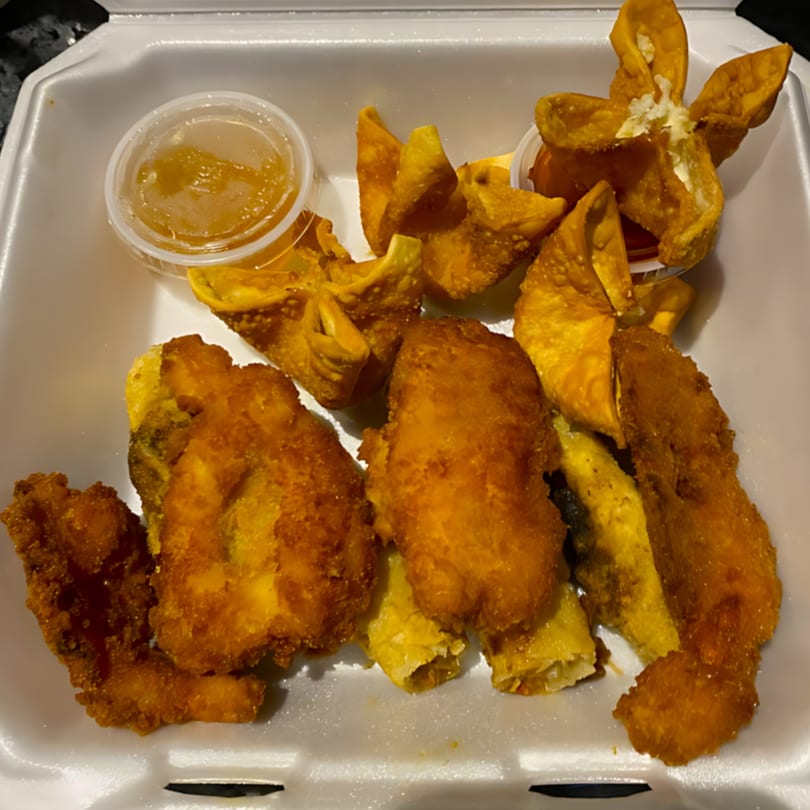 A takeout container filled with golden-fried chicken, spring rolls, and crab rangoons with dipping sauces.