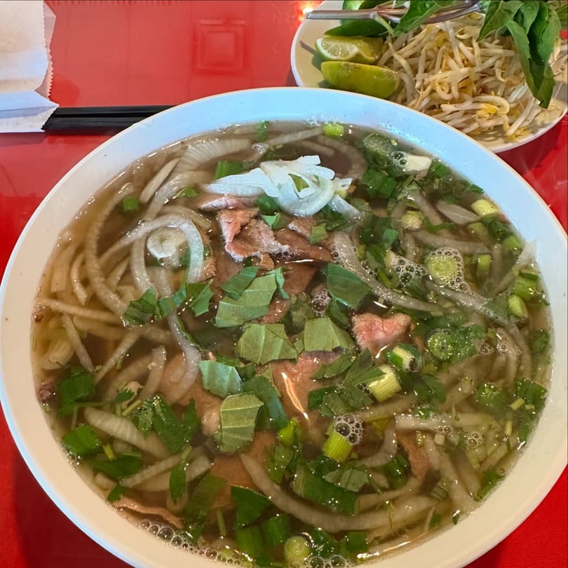 A large bowl of Vietnamese pho with beef, noodles, onions, and herbs, with a side of bean sprouts and lime.