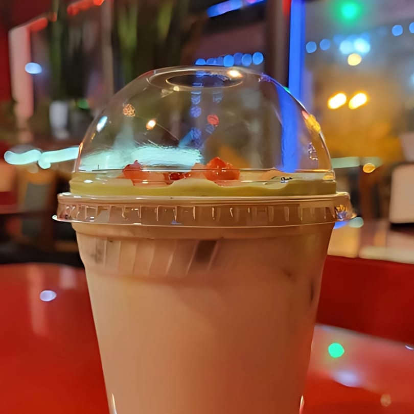 A Vietnamese drink with fruit toppings in a plastic cup, on a red table with blurred lights.